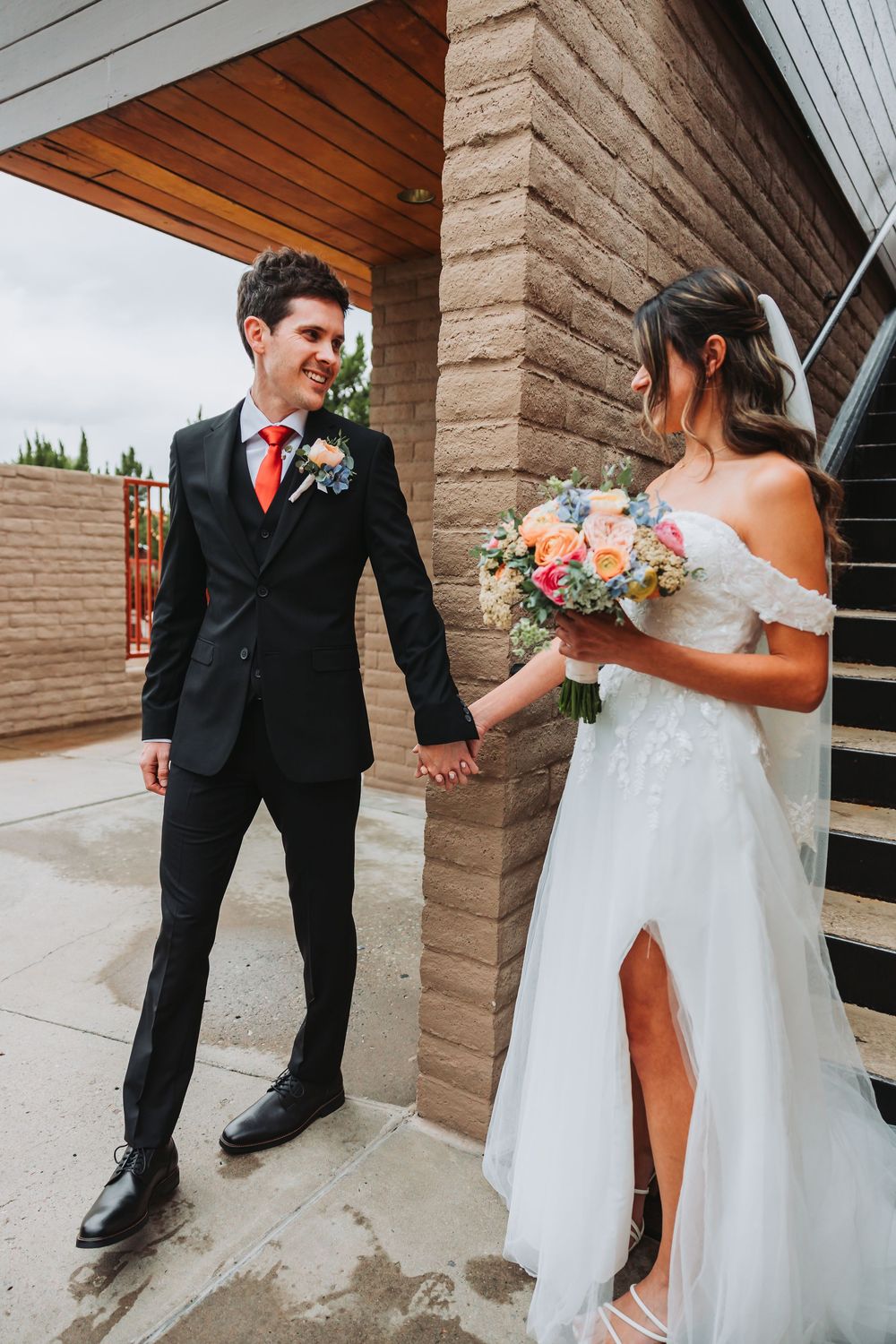 A couple on their wedding day holding hands, with the bride in white dress holding a peach bouquet and groom in black suit.