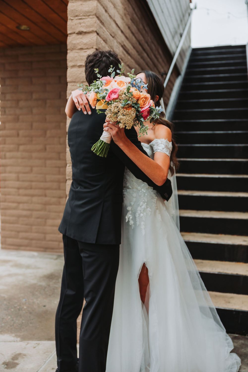 A bride and groom embrace while holding a vibrant peach and pink wedding bouquet near a staircase.