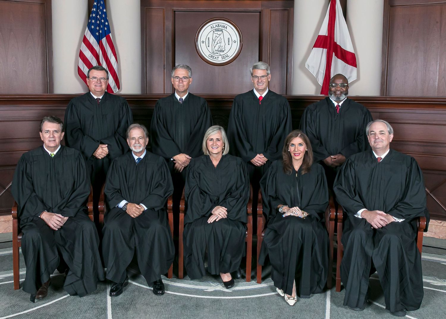 Nine judges in black robes seated in a formal courthouse setting with American and Alabama state flags displayed.