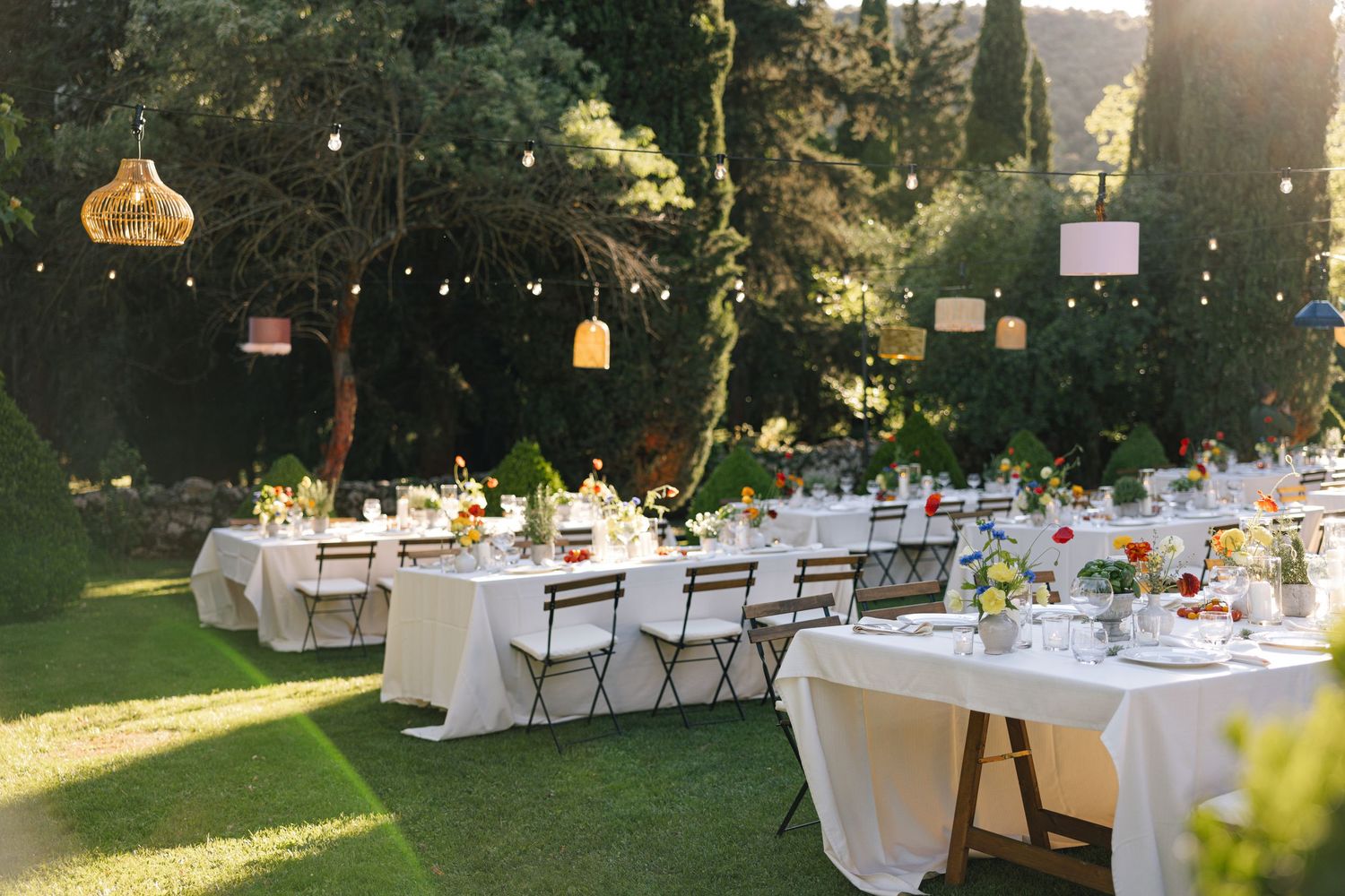 Elegant outdoor wedding reception setup with white-clothed tables and chairs arranged in a garden setting at sunset.