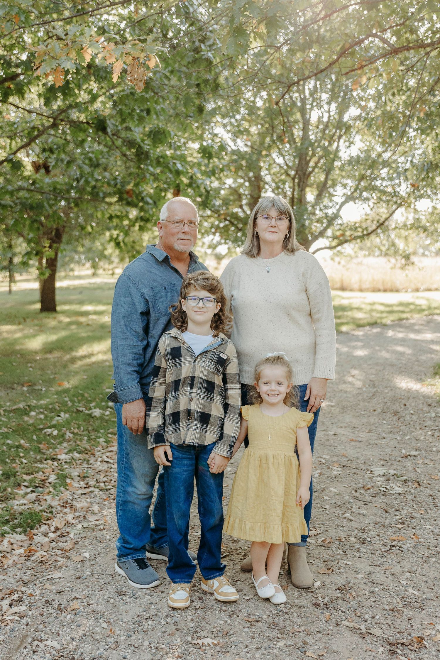 A family of four poses together on a dirt path surrounded by autumn trees in a park setting.