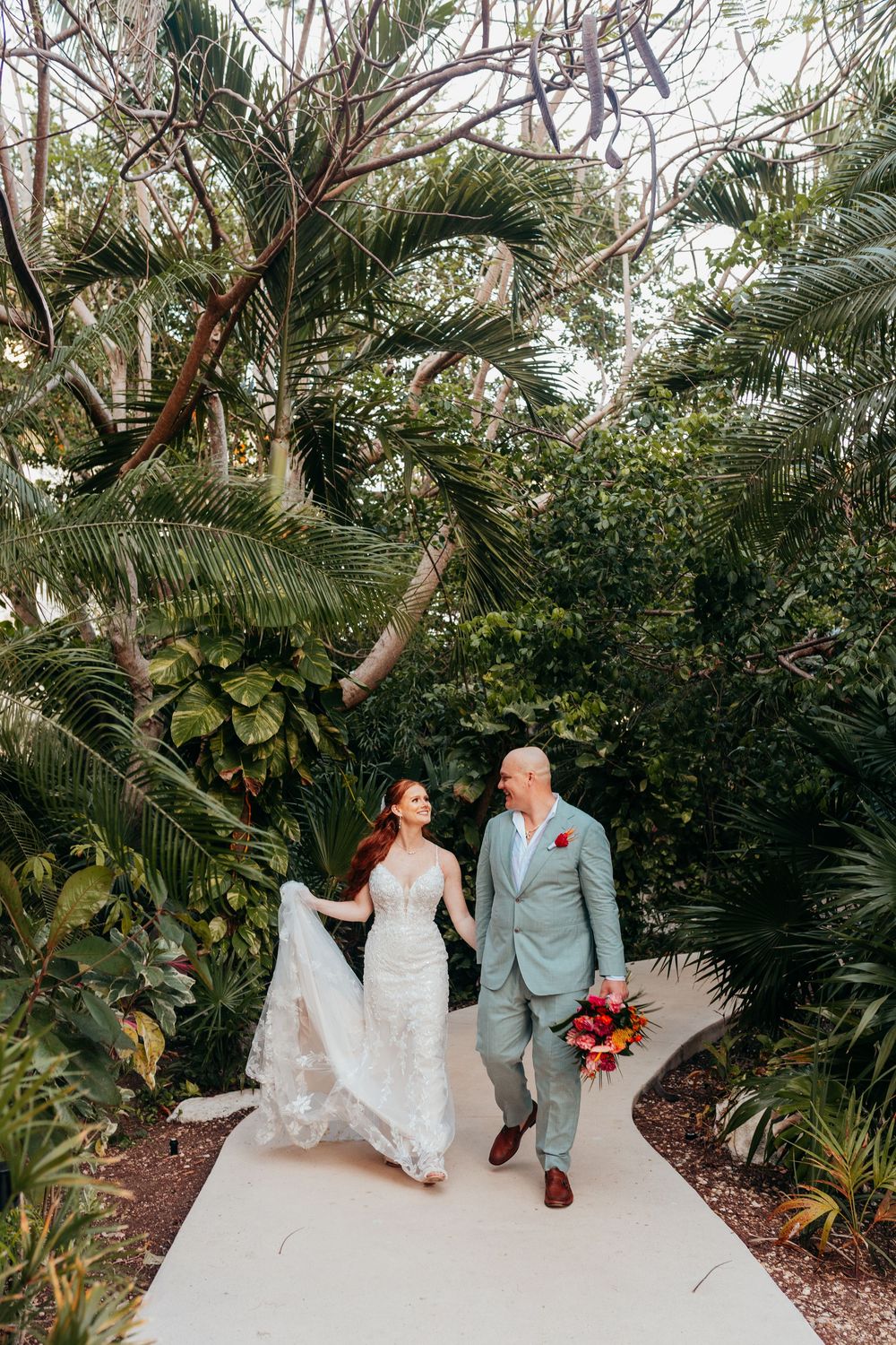 Newlywed couple walks hand in hand along tropical garden path surrounded by lush palm trees and greenery.