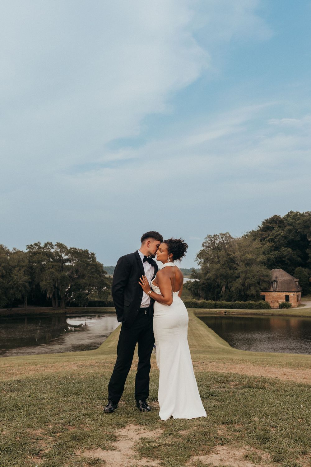 A couple in formal attire shares a romantic moment by a serene lake with trees in the background.