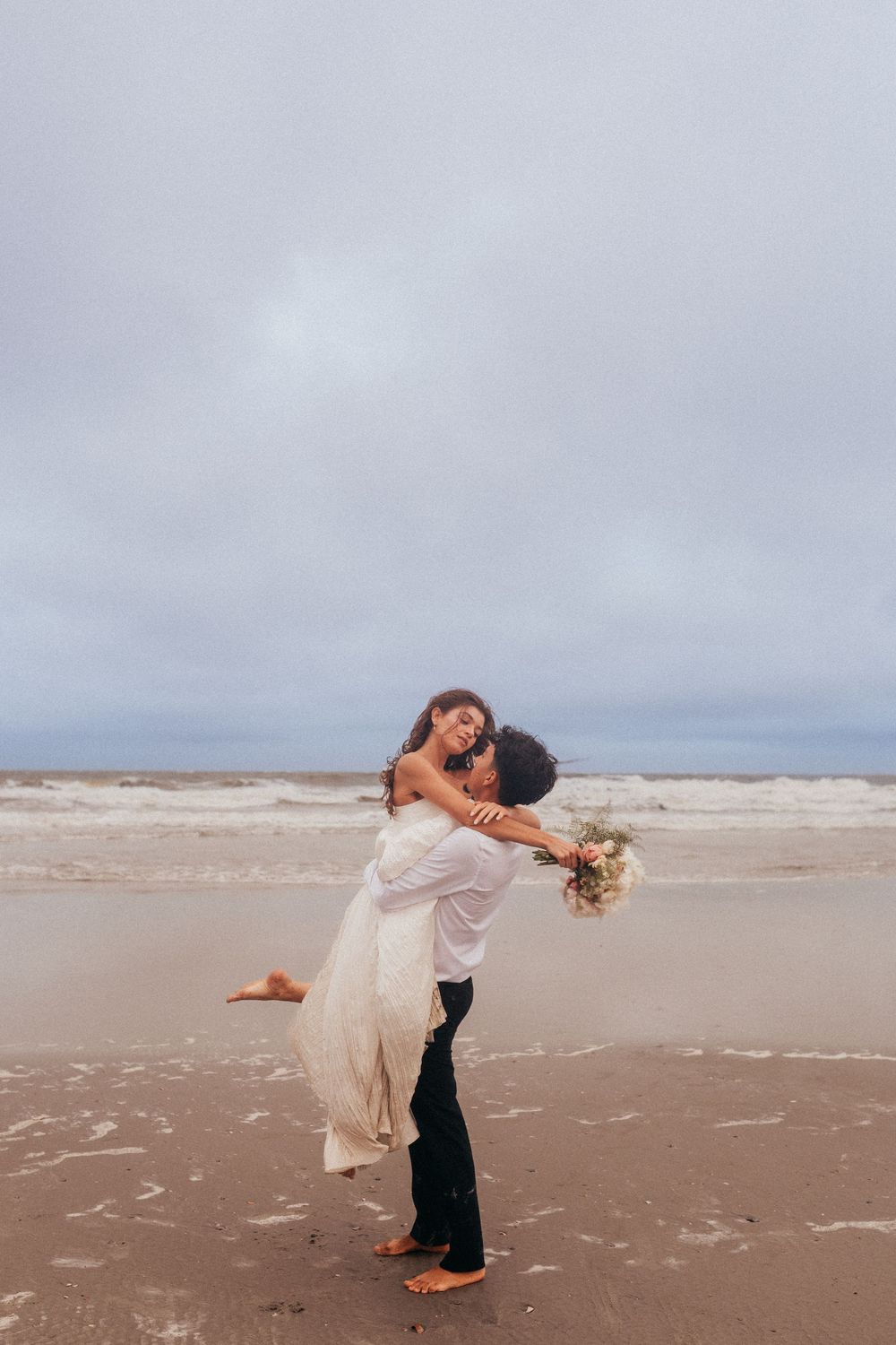 A romantic couple embraces on a cloudy beach, with the bride in a flowing white dress holding a bouquet.