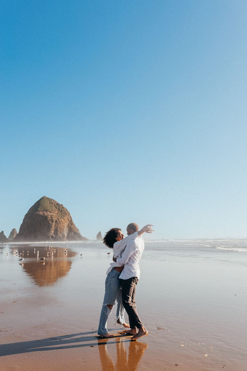 Couple embraces on sandy beach with Haystack Rock at Cannon Beach, Oregon in the background under clear blue skies.