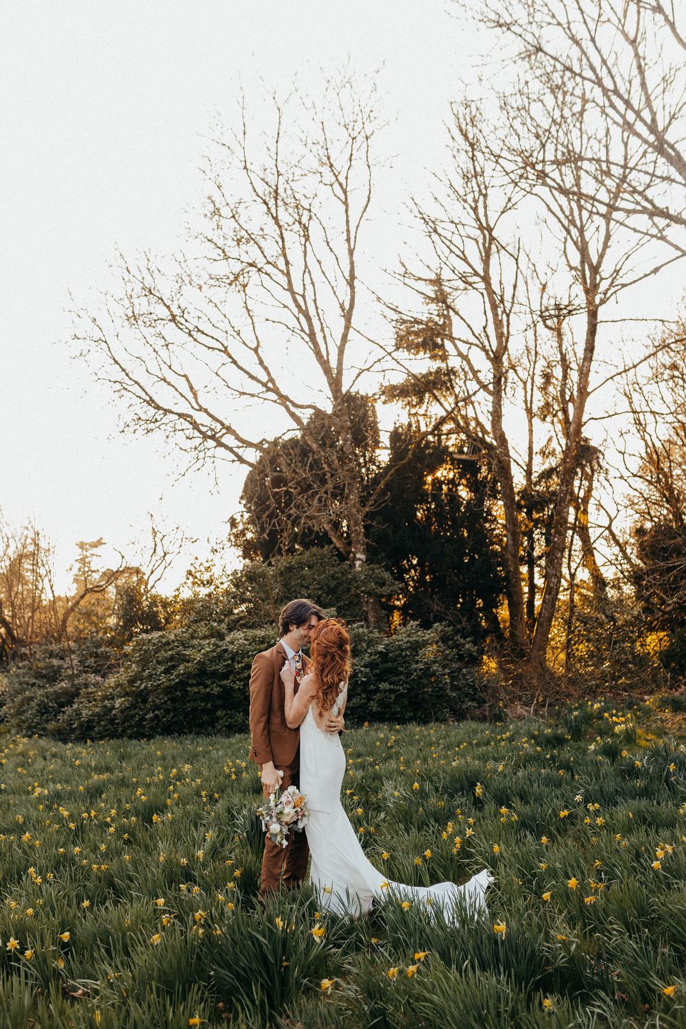 A couple in wedding attire share an intimate moment together in a wildflower meadow at sunset.