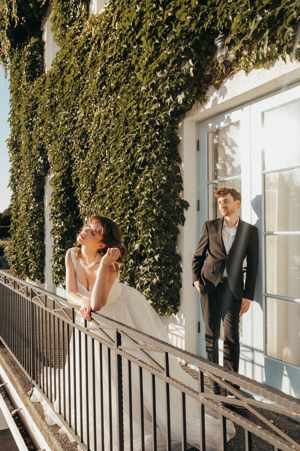 Two people in wedding attire pose on a balcony with lush green ivy growing up the white wall behind them.