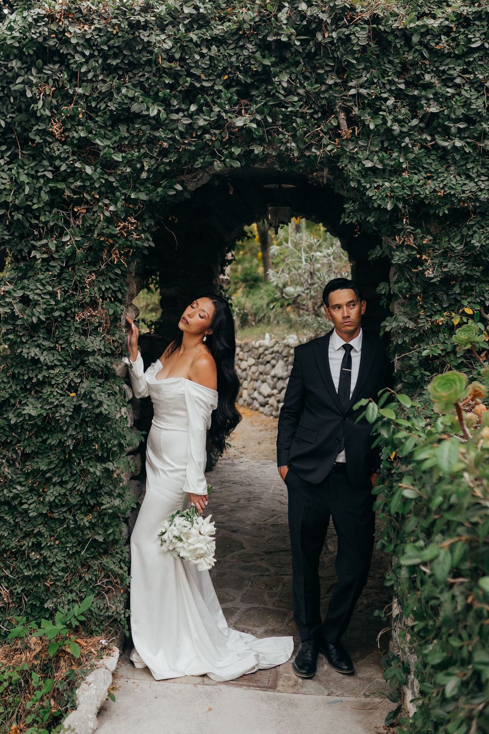 Elegant couple walks through lush garden pathway in romantic wedding photoshoot series.