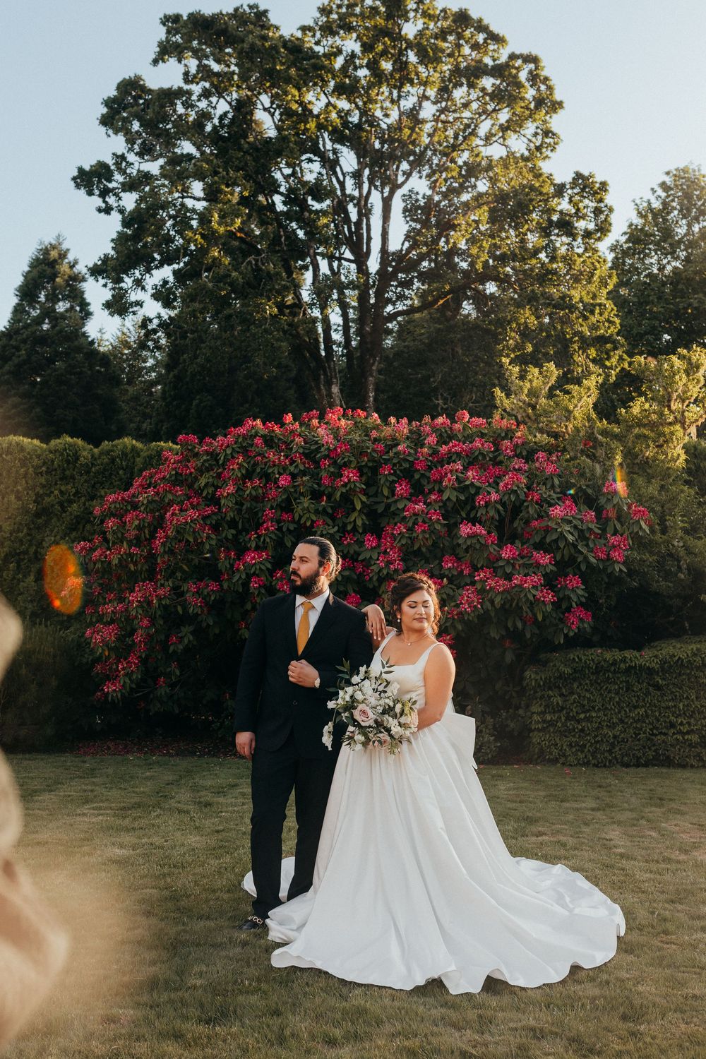 A couple stands in front of a vibrant pink azalea bush during golden hour at an outdoor garden wedding ceremony.