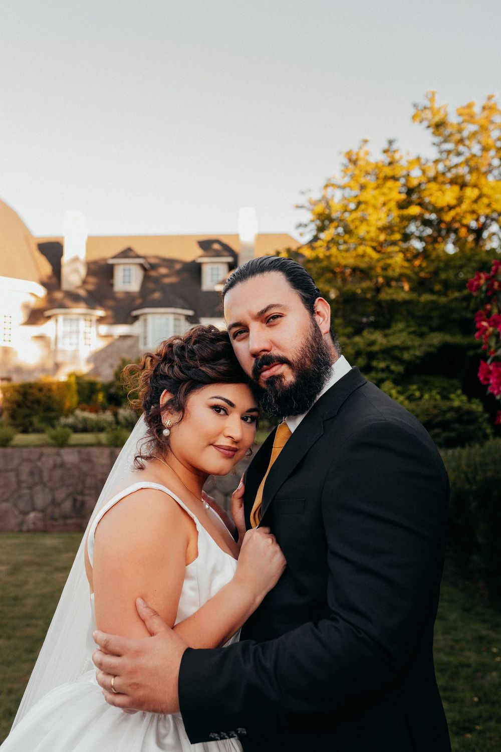 Romantic wedding couple embrace outdoors during golden hour with autumn trees and buildings in background.
