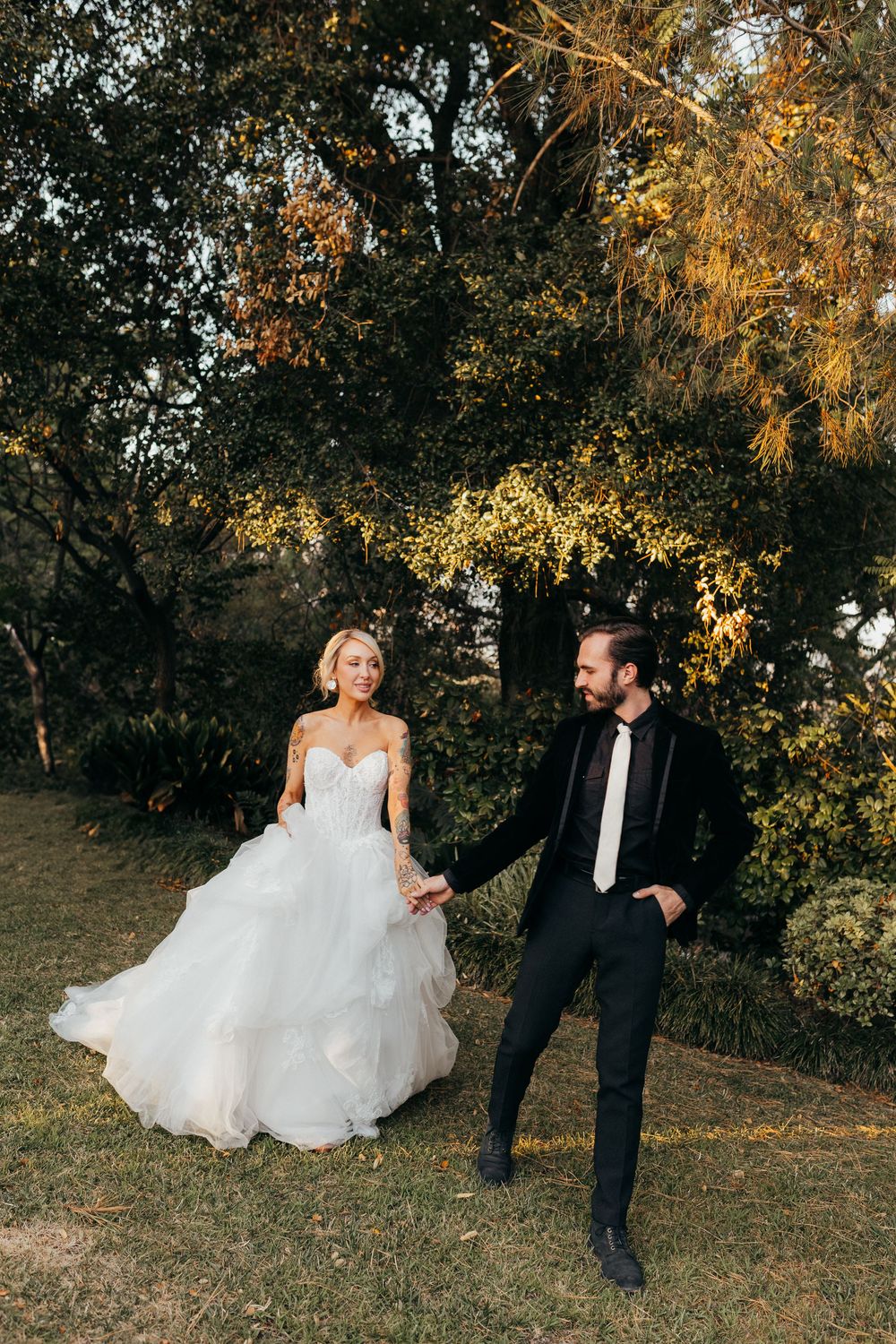 A couple in wedding attire hold hands while standing in a sunlit outdoor setting with autumn trees in the background.