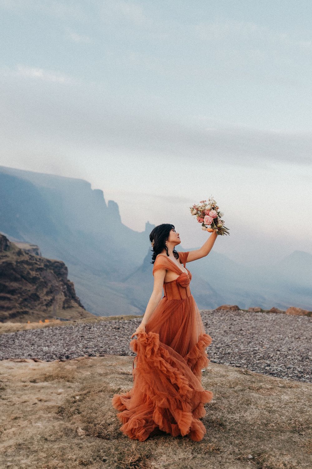A woman in a flowing rust-colored dress holds up a bouquet against a dramatic mountain landscape at sunset.