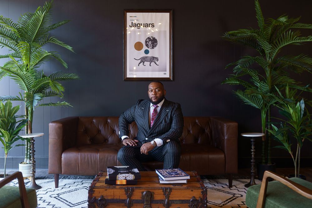 A modern office living room features a brown leather couch, potted plants, and decorative wall art against a dark wall.