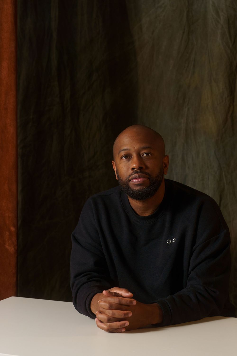 A person wearing a black sweater sits at a white table against a dark textured background.