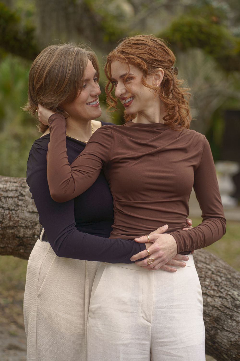 Two people in brown and black tops with white pants embrace while laughing together on a stone surface outdoors.