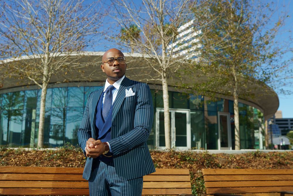 Professional in a pinstriped suit stands outdoors near modern office buildings and benches on a sunny day.