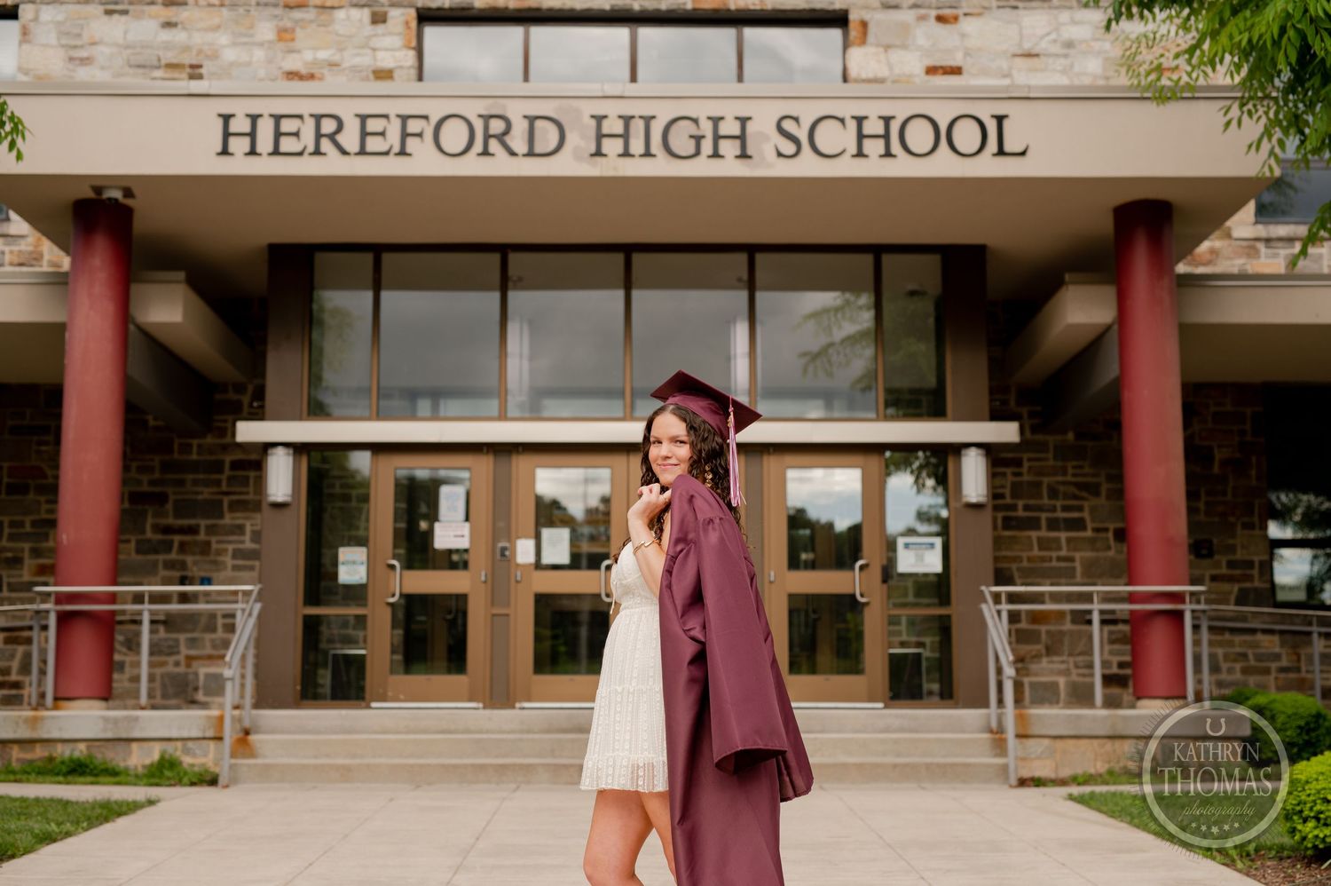 Graduate in white dress and burgundy cap poses in front of school columns.