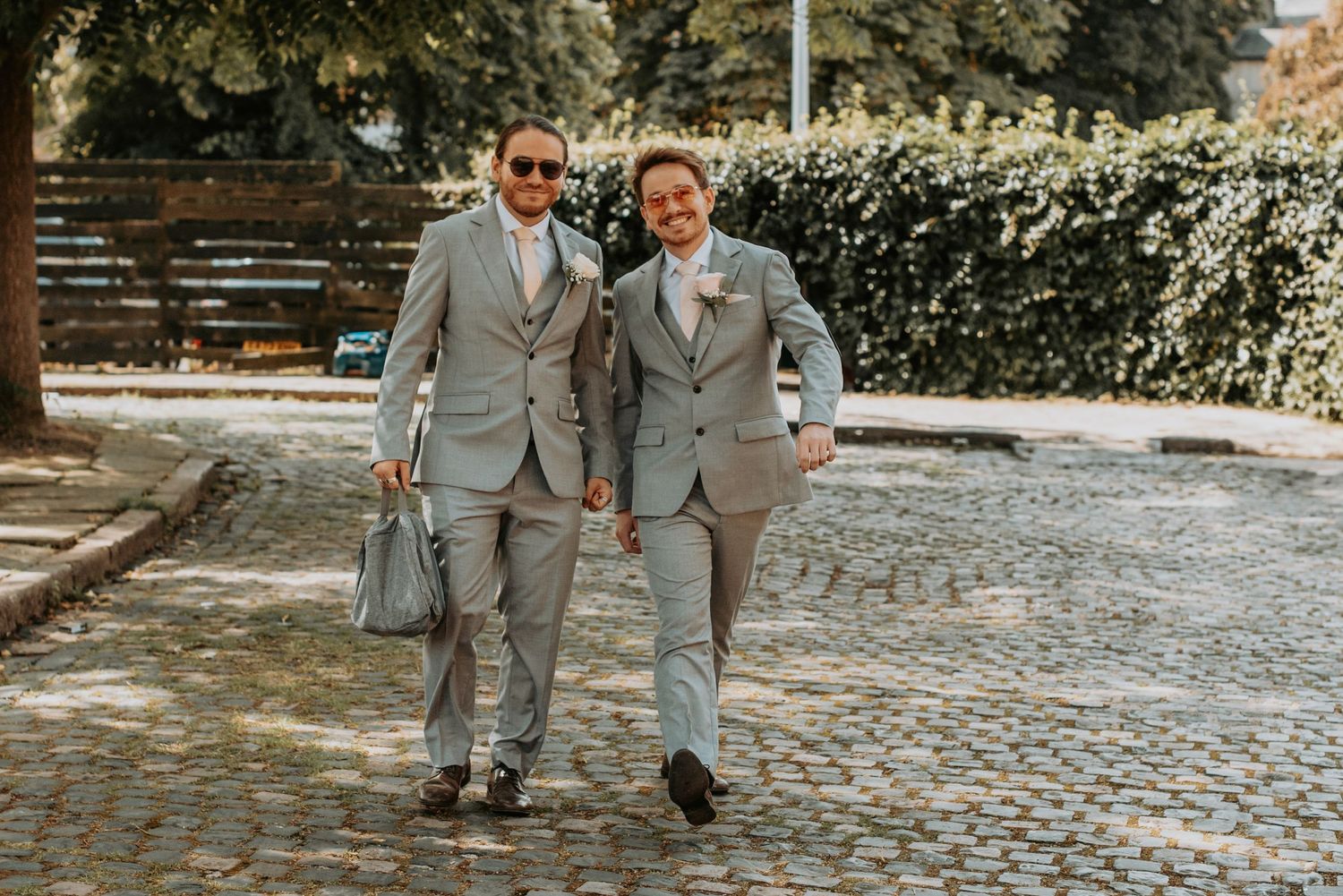 Two people in matching light gray suits walk together on a cobblestone path in a garden setting.