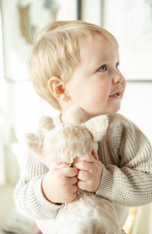 Toddler in cream knit sweater holding stuffed toy looks up with bright smile.