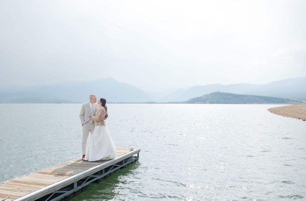 Two people in white outfits walk along a wooden pier extending into a tranquil lake surrounded by mountains.