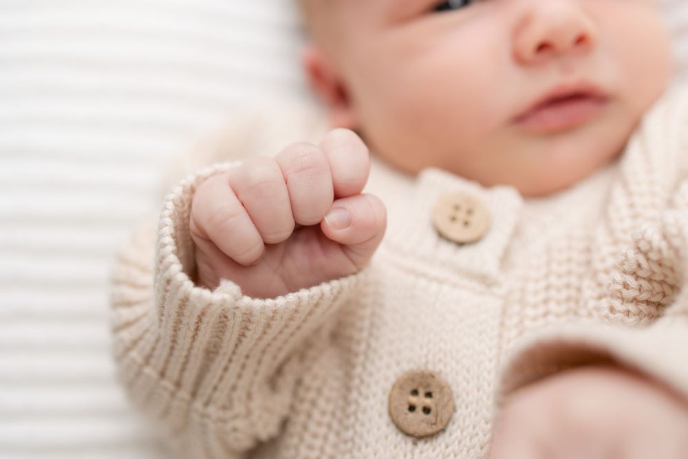 Baby's tiny hand reaches out while wearing a cozy cream-colored knit cardigan with wooden buttons.