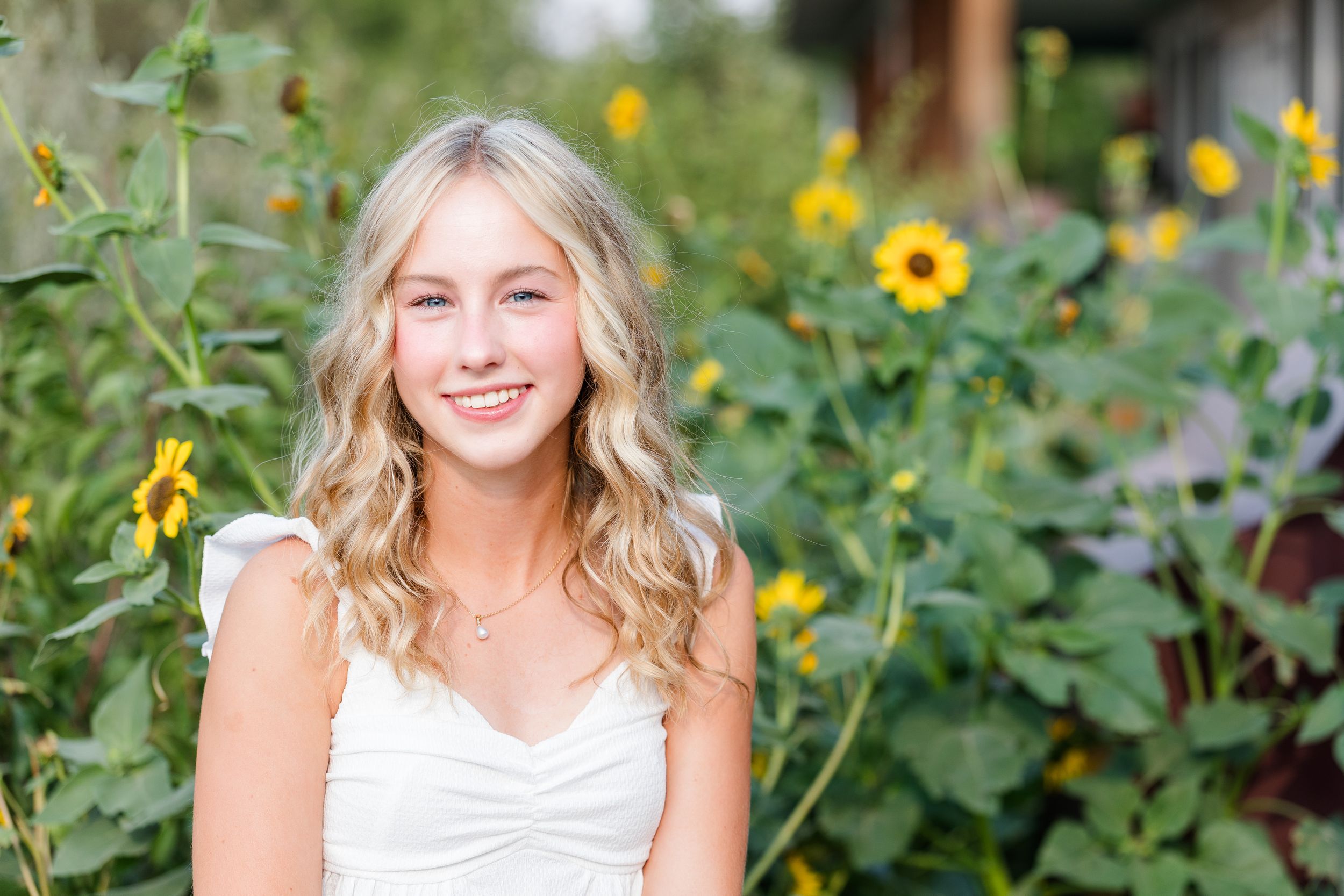 Smiling blonde teenage girl in white dress poses among yellow sunflowers in a sunny outdoor garden setting.