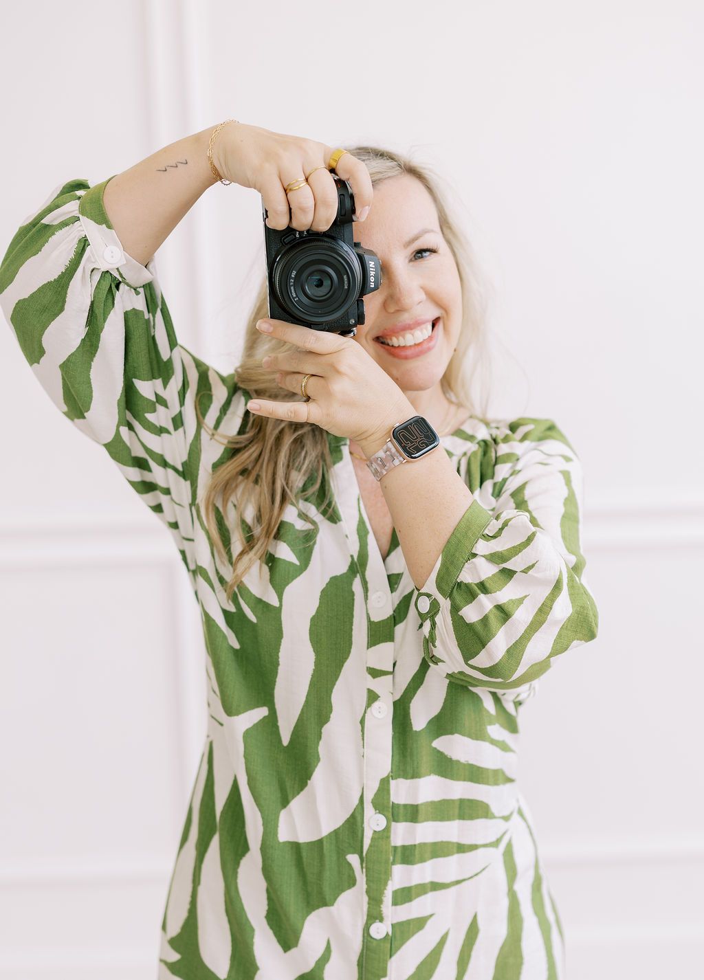 A female photographer in a white blouse holding a camera near large windows in a bright indoor space.