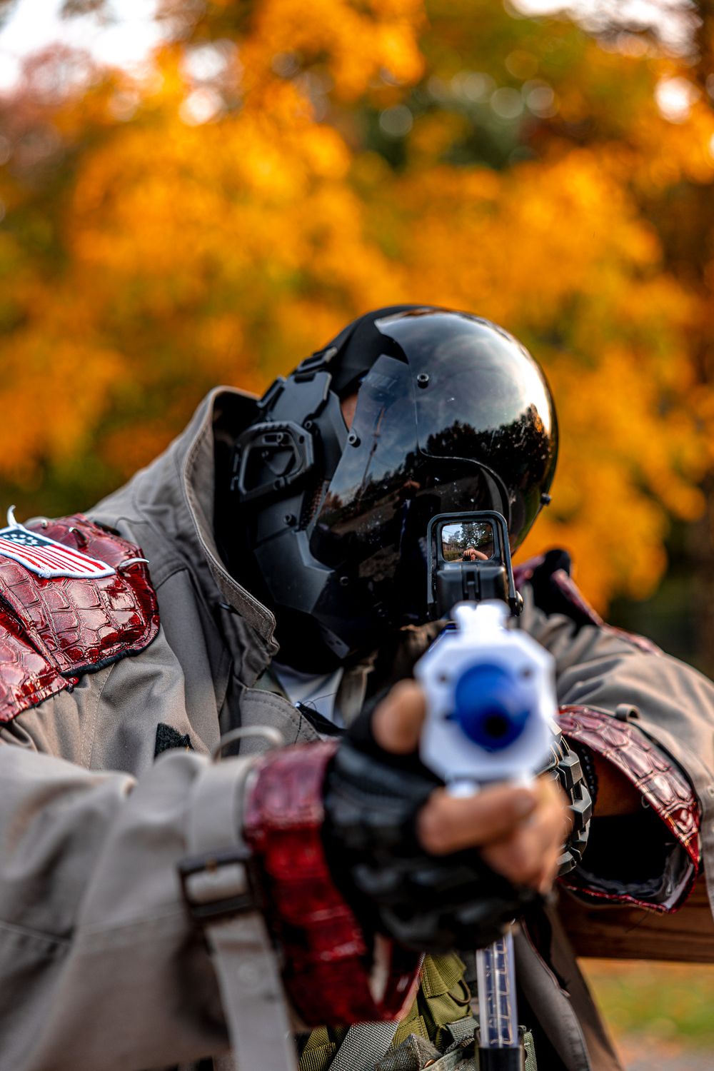 A paintball player in black mask aiming with blue marker against autumn foliage background.
