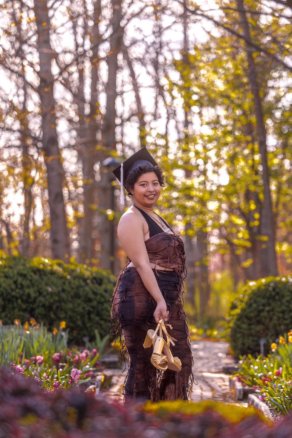 A ballet dancer stands among autumn flowers and trees in a garden while holding pointe shoes at sunset.