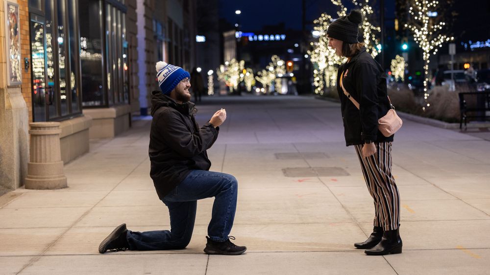 A romantic evening proposal on an illuminated city street with holiday lights and decorations.