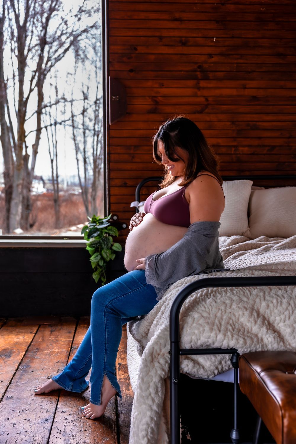 An expecting mother in purple and jeans sits contemplatively by a window in a rustic bedroom with wood walls and white bedding.