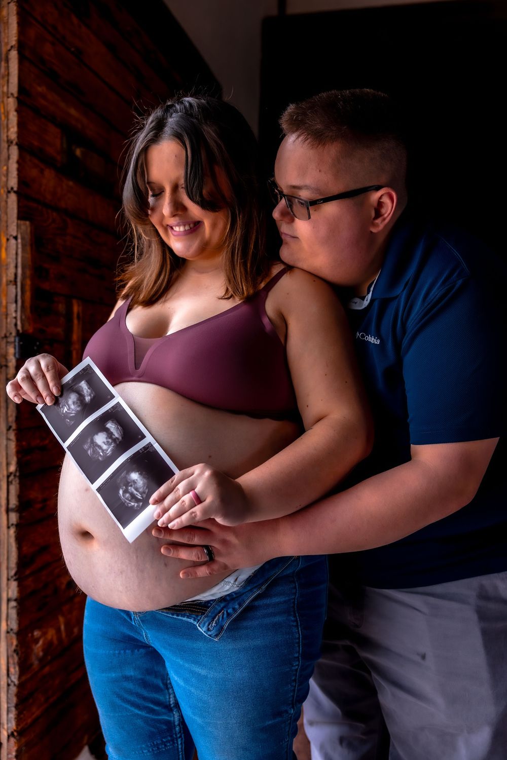 Expectant couple holding ultrasound photos against a rustic brick wall background.