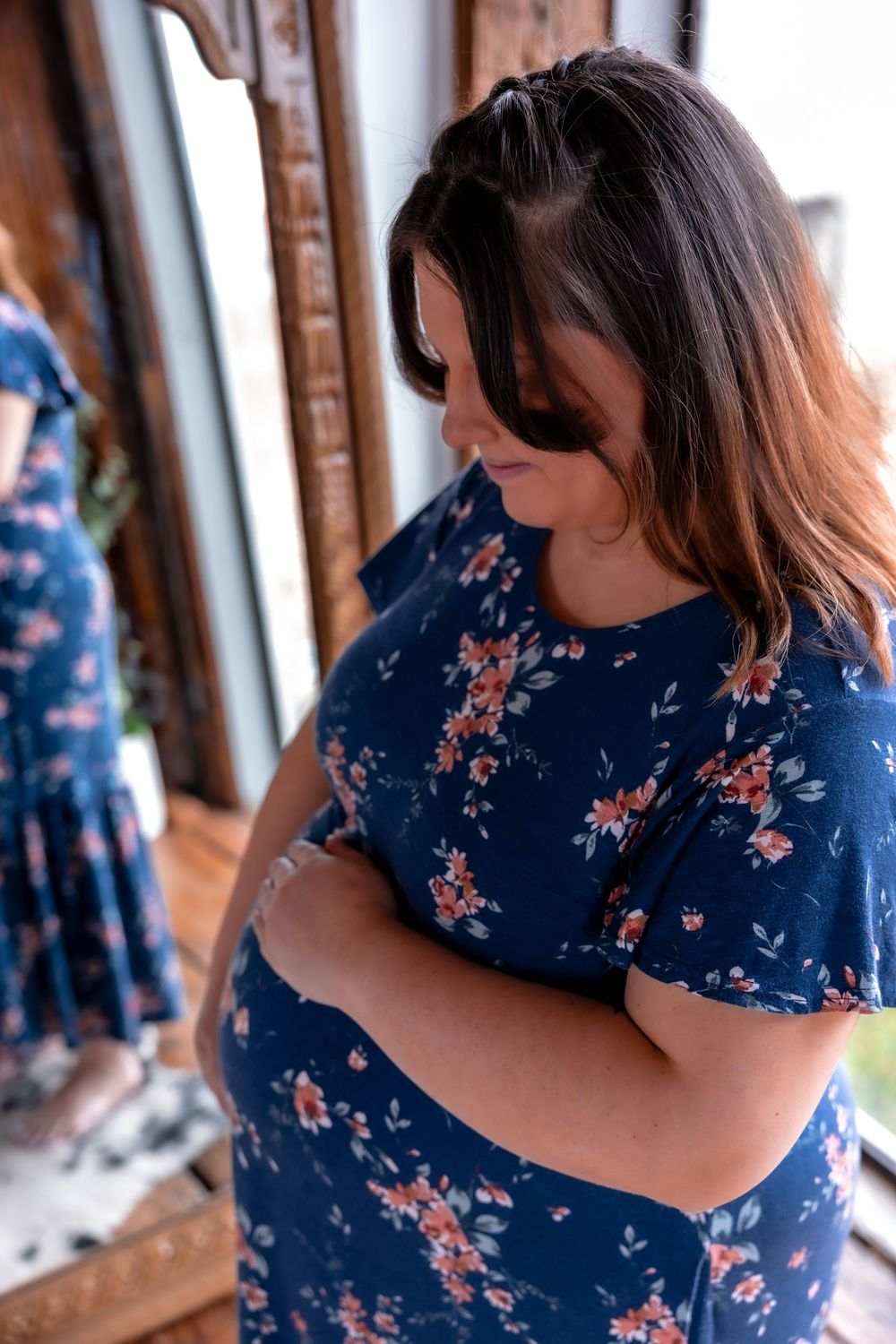 Pregnant woman in a blue floral dress standing near a window with natural light.