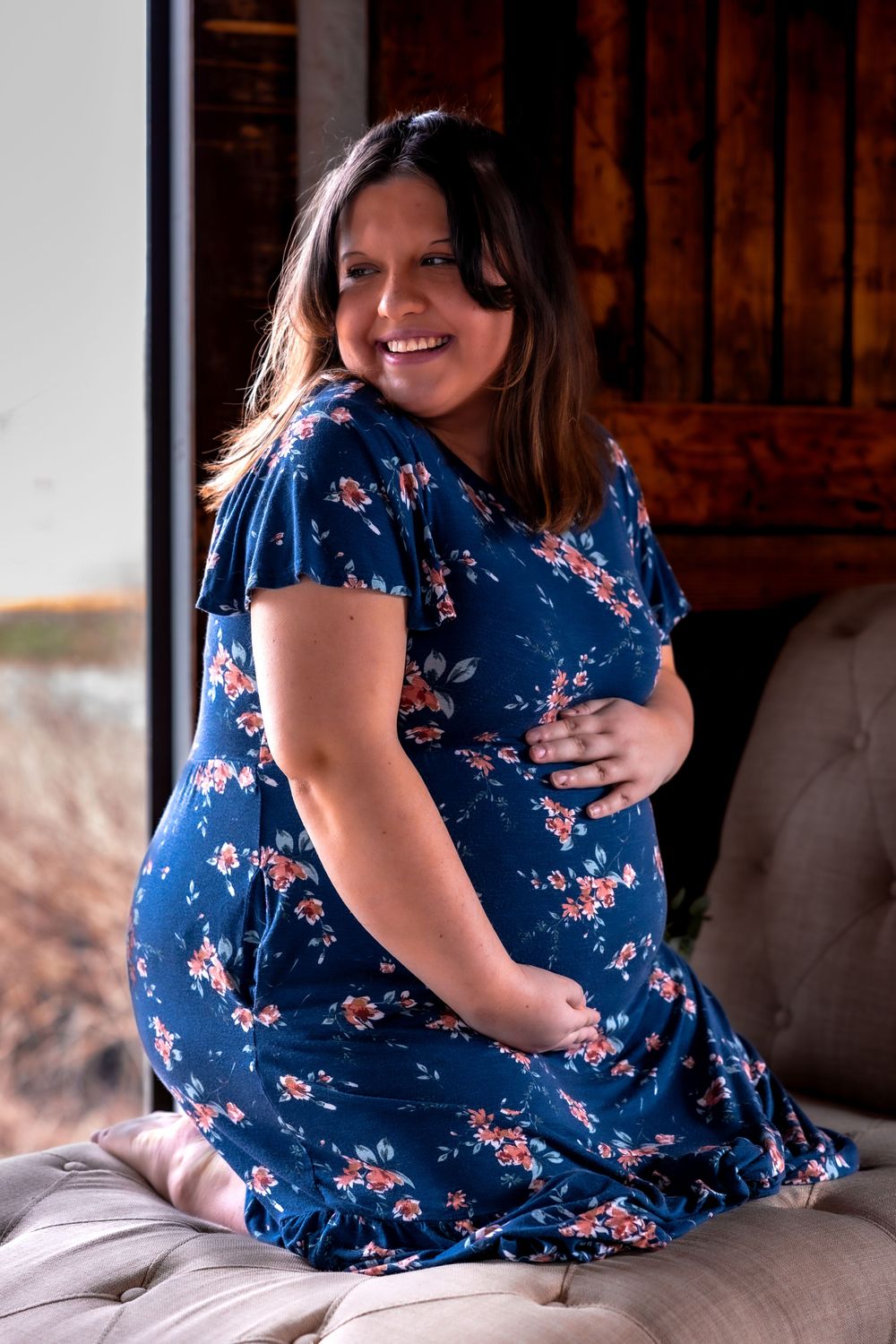 Smiling pregnant woman in a navy floral dress sitting on cushioned surface.