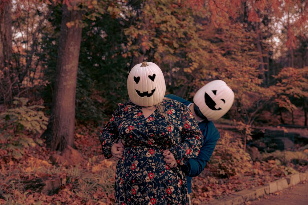 Two people wearing pumpkin head masks and autumn clothing pose in a forest with orange fall foliage in the background.