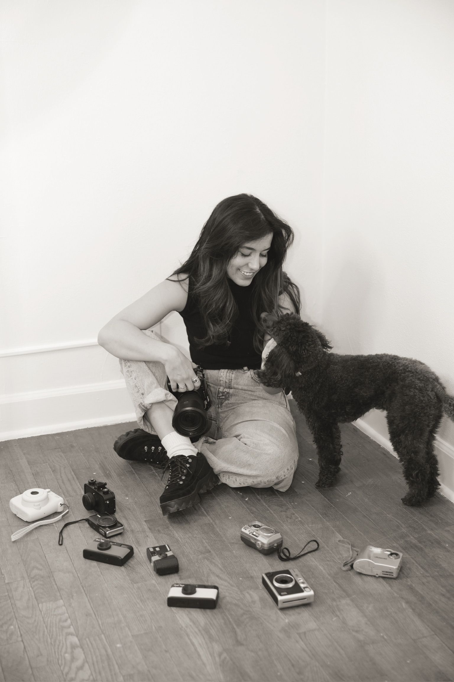 A person sits on hardwood floor with a black poodle while various vintage cameras and equipment are scattered around.