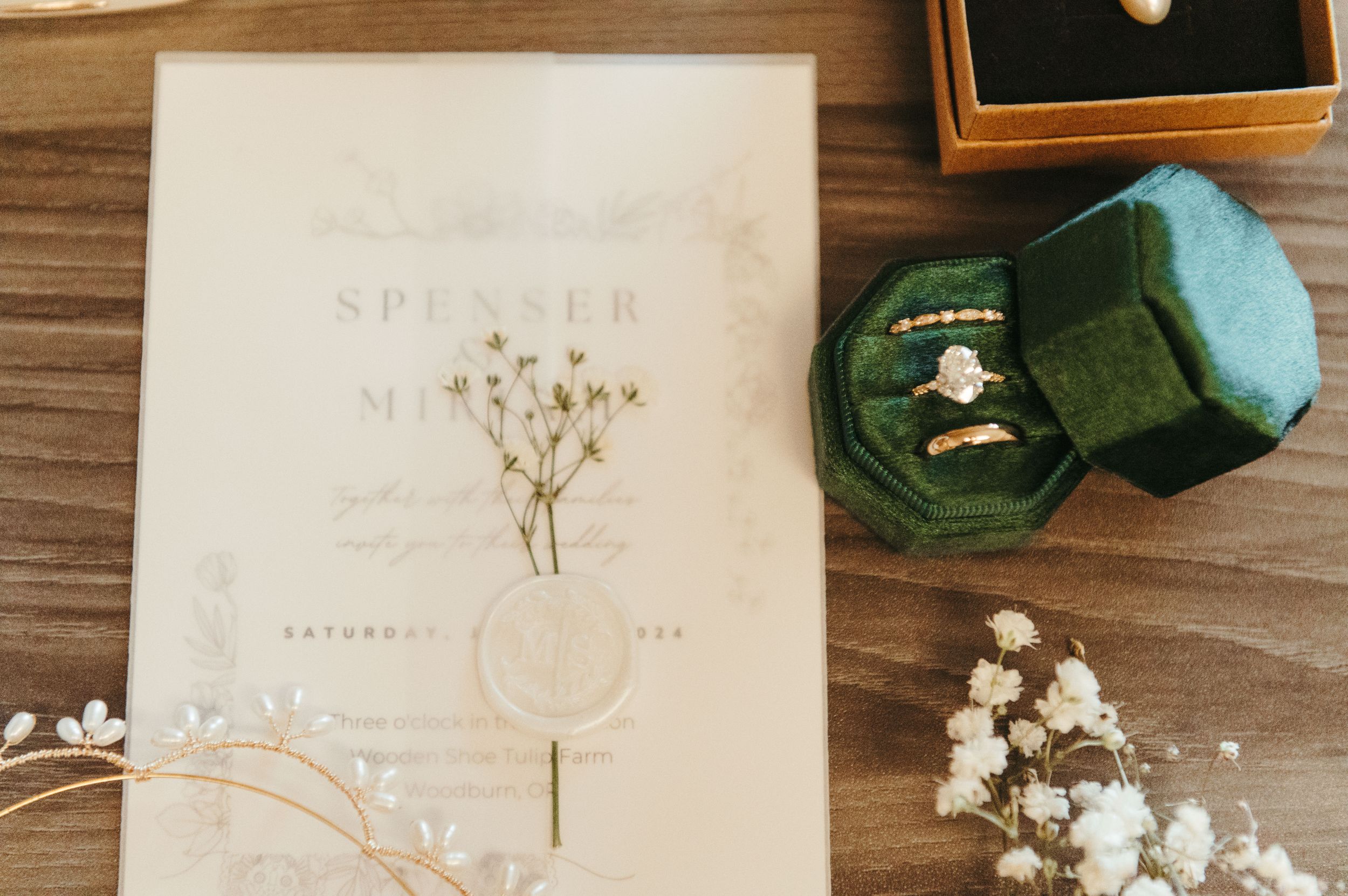 Wedding invitation with gold lettering next to a green velvet ring box and delicate baby's breath flowers on wooden surface.