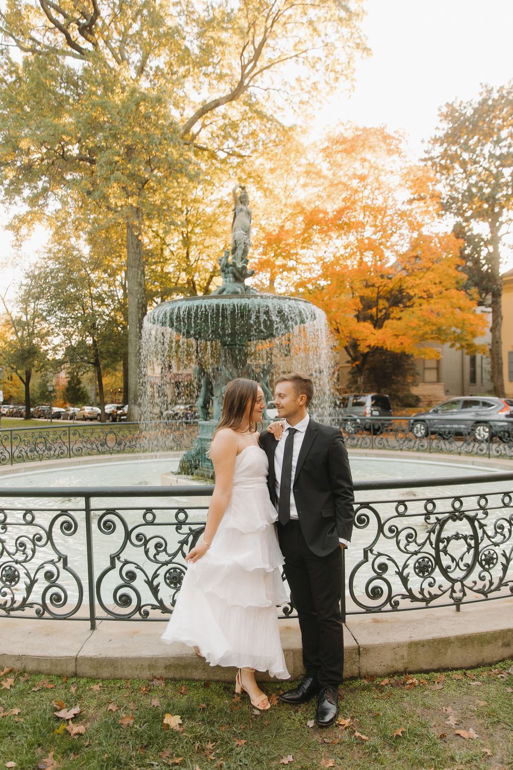 A couple in wedding attire sits together on a decorative stone wall surrounded by autumn foliage in a park setting.