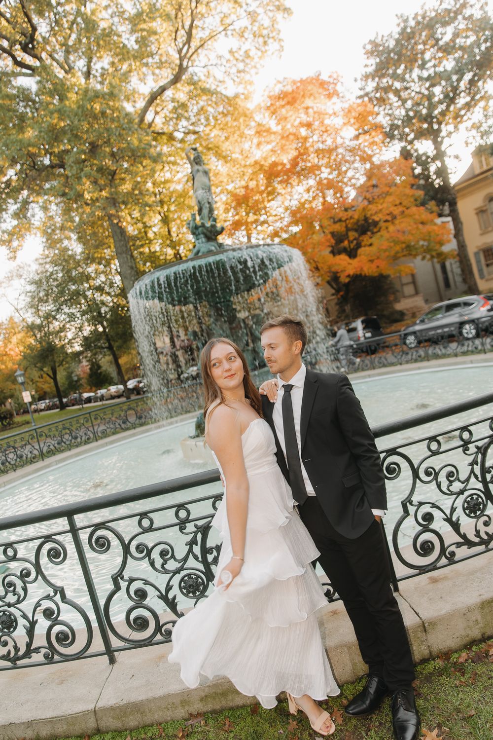 A couple in wedding attire poses together on a wrought iron bridge surrounded by autumn foliage in a park setting.