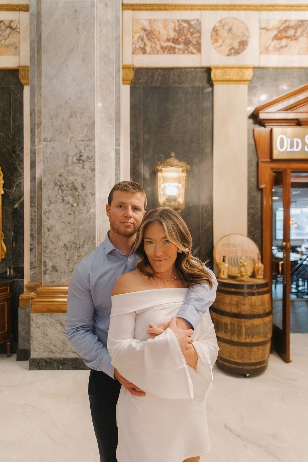A couple poses together in an elegant indoor setting with marble columns and wooden barrels in the background.