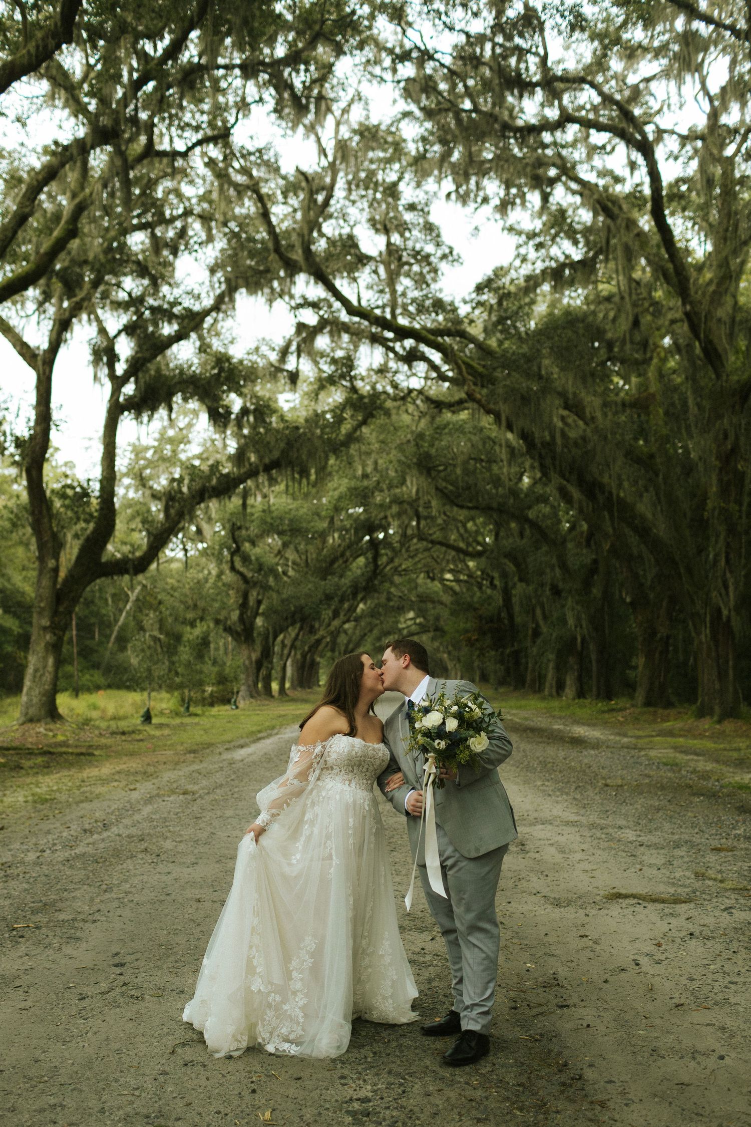 A bride and groom kiss at the end of a long tunnel of trees covered in Spanish moss in Savannah, GA