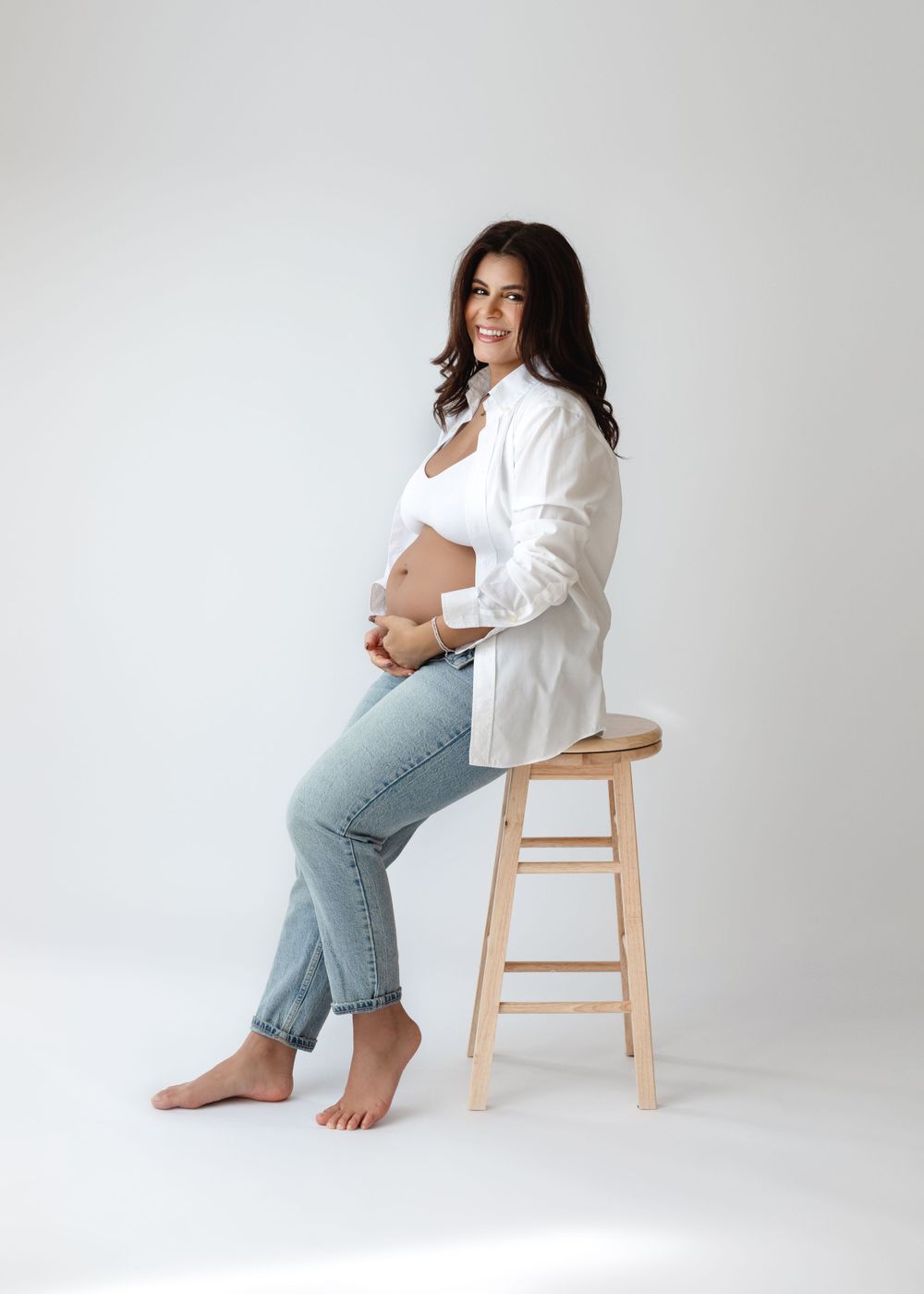 Maternity portrait of woman in casual attire sitting on wooden stool against white backdrop.