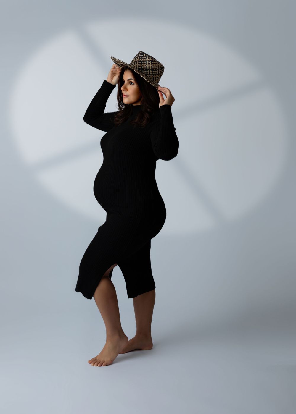 Stylish maternity pose in black dress and plaid hat against grey backdrop.