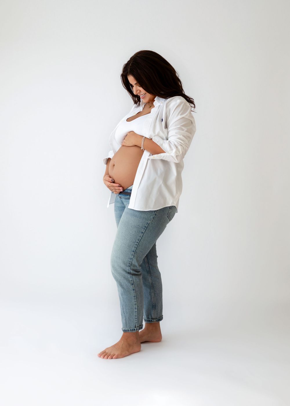 Side view of expectant mother in white shirt and jeans touching her belly against white background.