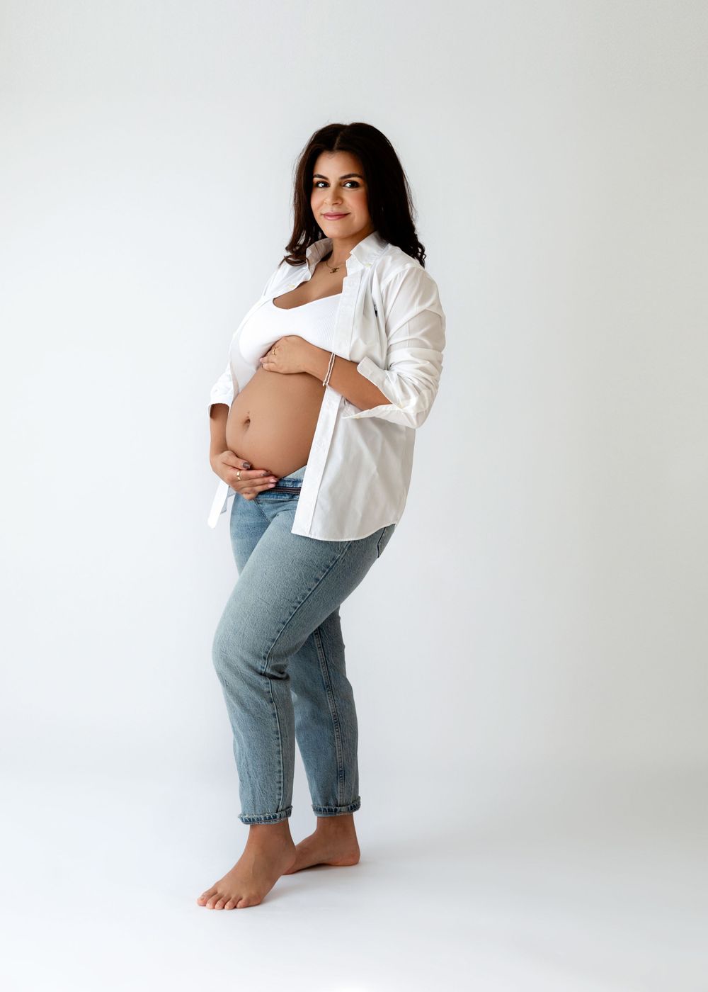 Pregnancy portrait showing woman in white shirt and jeans posing against white background.