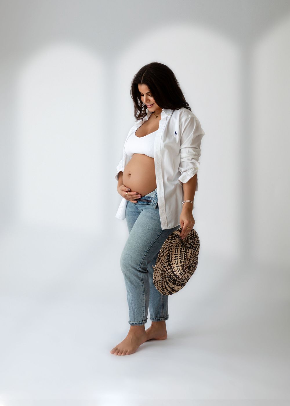 Expectant mother in casual white shirt and jeans poses against white background holding woven bag.