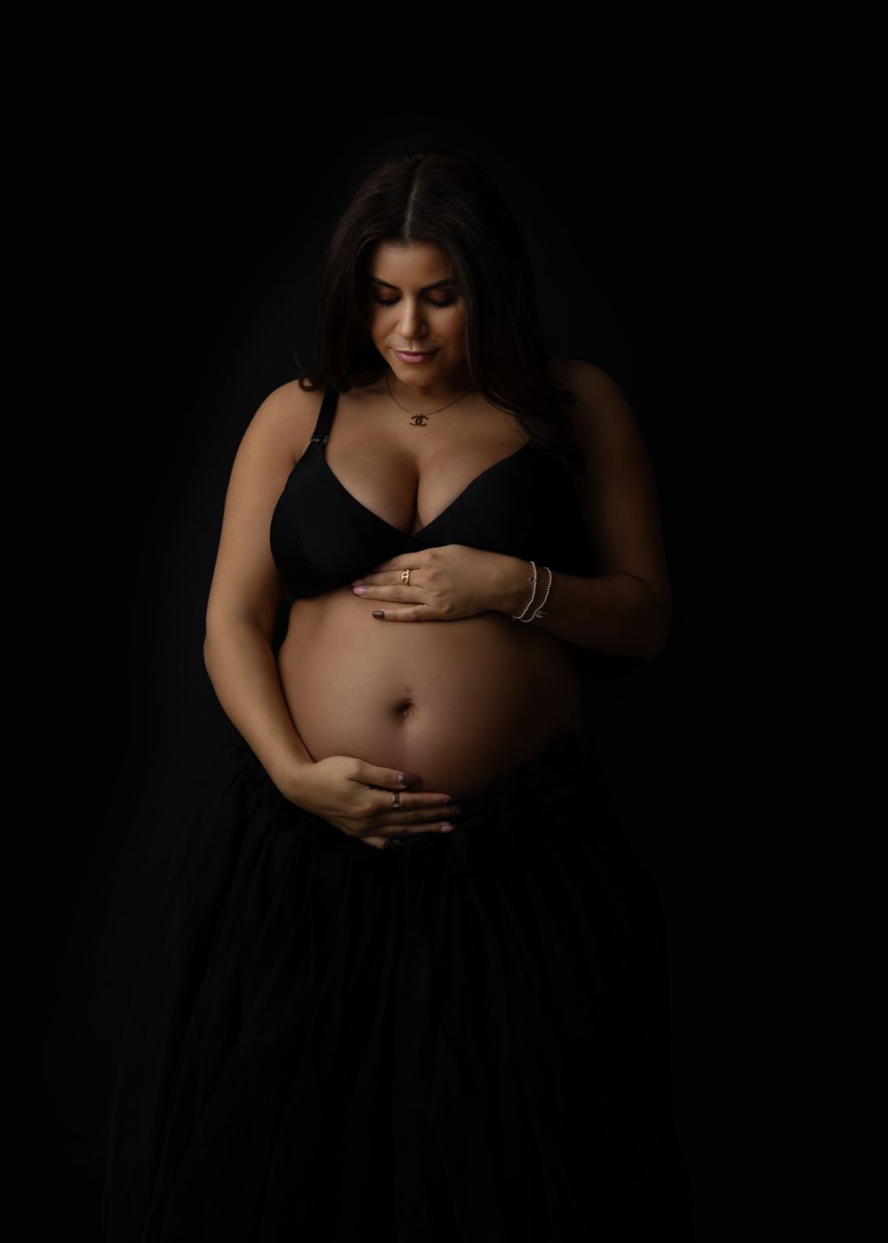 A maternity portrait against black background shows an expecting mother in black top cradling her belly.