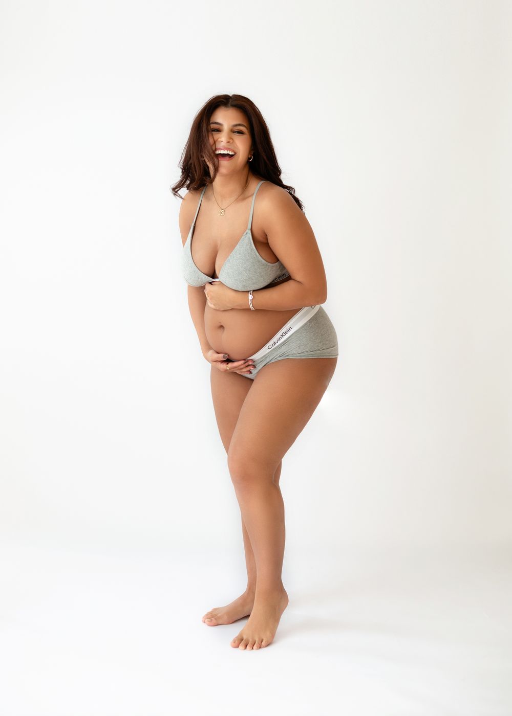 Confident woman in grey lingerie set poses playfully against white studio backdrop.