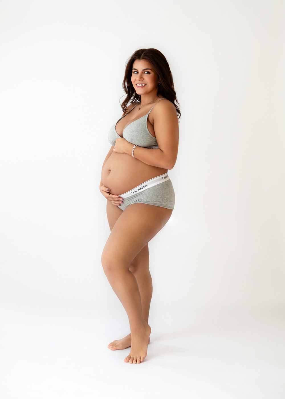 Pregnant woman in grey underwear set poses elegantly against minimalist white background.