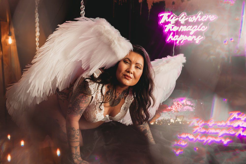 Someone in white angel wings poses on bed with pink neon sign glowing in background during a boudoir photoshoot.